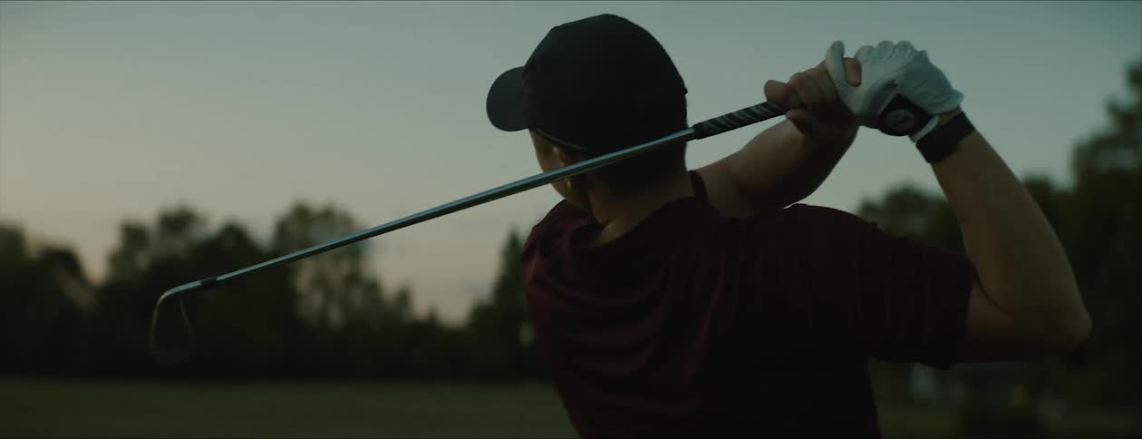 A man swings a golf club on a golfing green in the evening