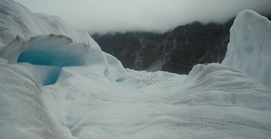 Blue interior of ice cave contrasts with ice and mountains in the background under an overcast sky
