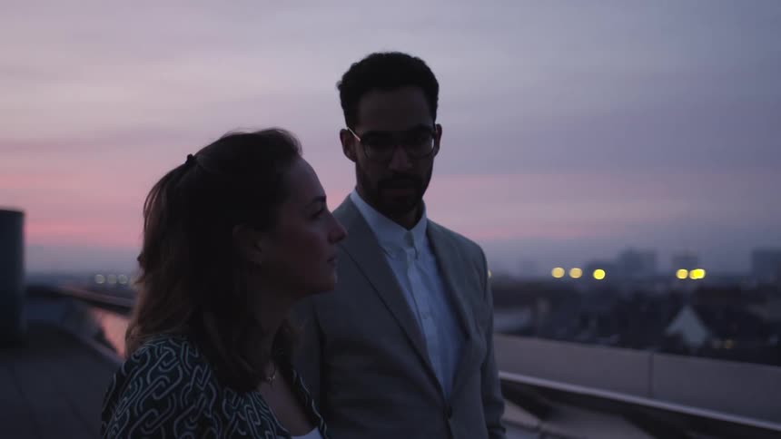 Man points while talking to woman as they stand on roof in city under a red sky