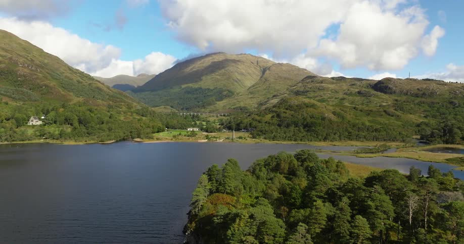 Lone vehicle moves along road near home nestled between lake and mountain, Glenfinnan Scotland