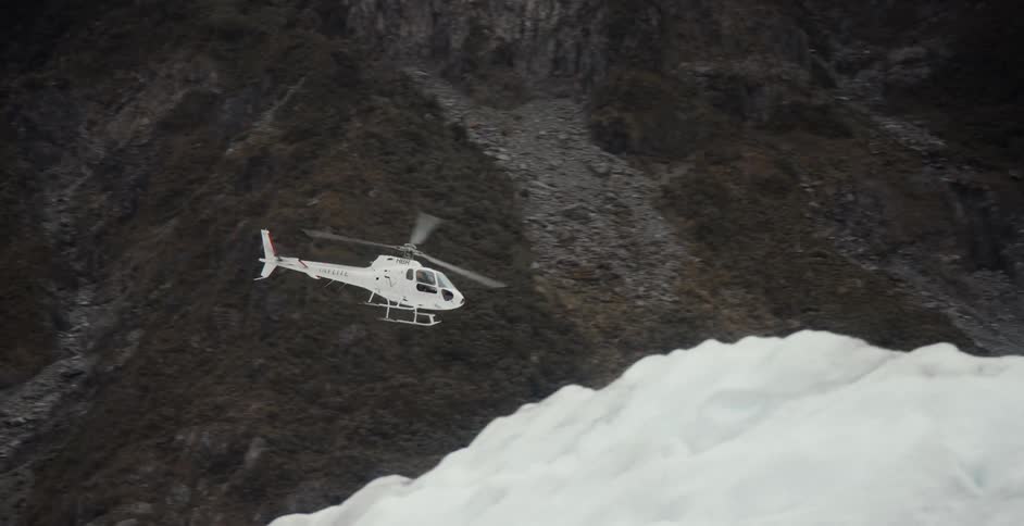 White helicopter flies over snow at base of mountain under low lying clouds