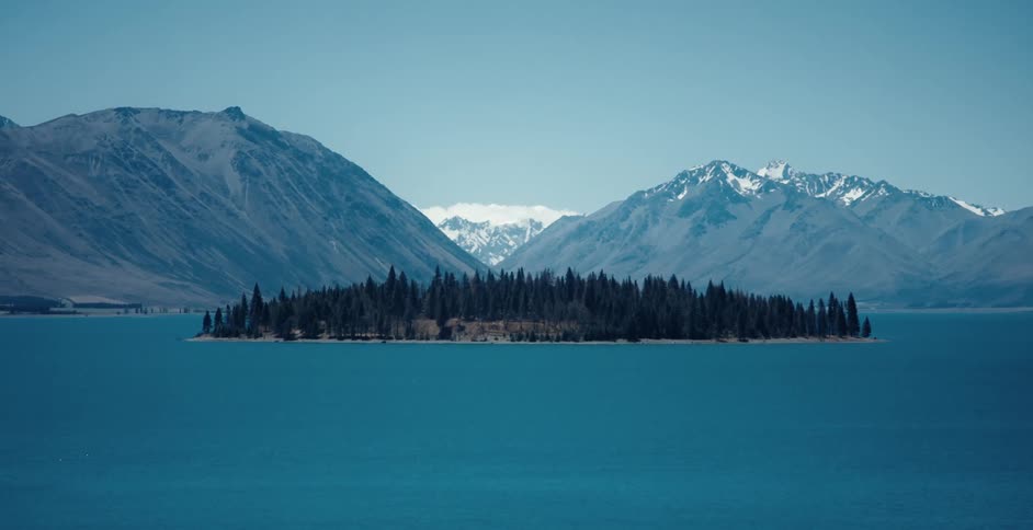A forest covers a small island near snow covered mountains