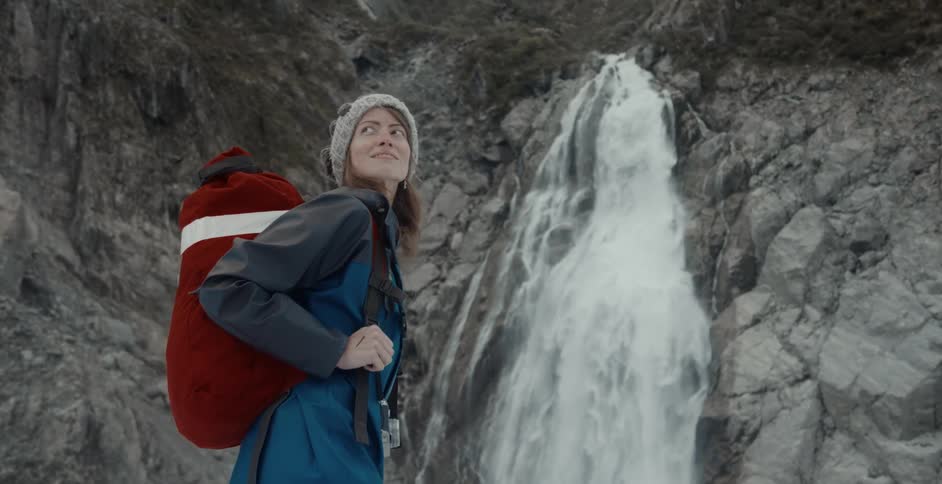 Female hiker with red backpack stands looking around near flowing waterfall