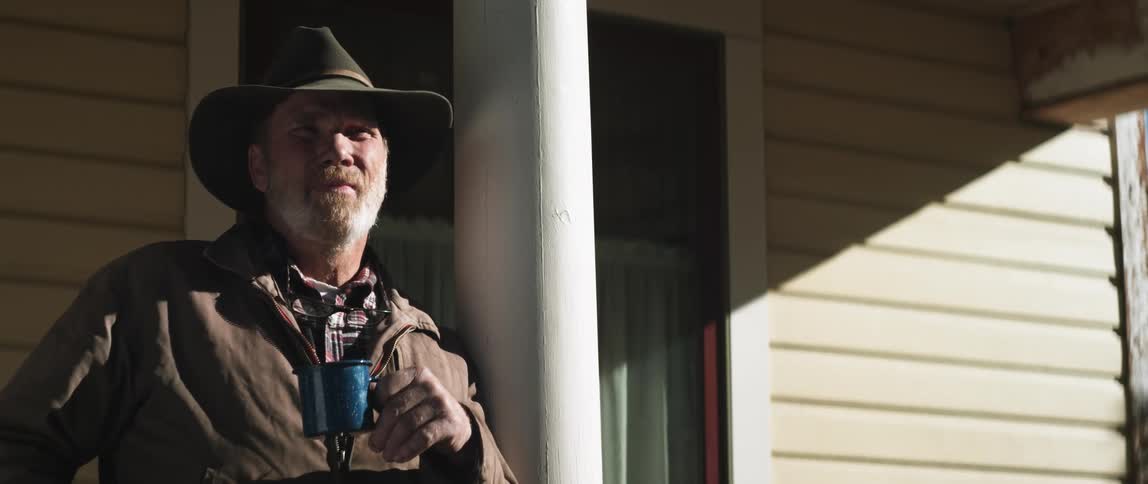 A man is leaning on a post outside a building and drinking coffee from a mug