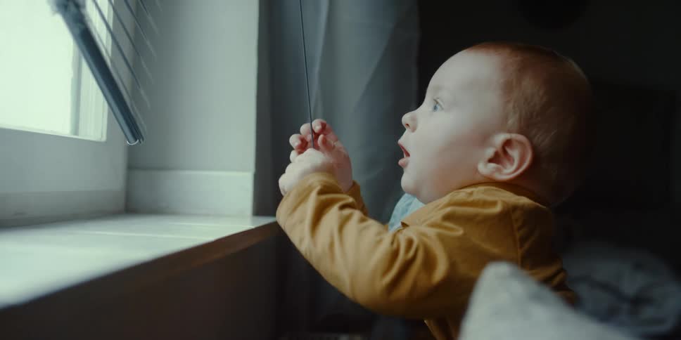 A young boy plays with a cord on blinds at home and waves his arms up when the blind moves