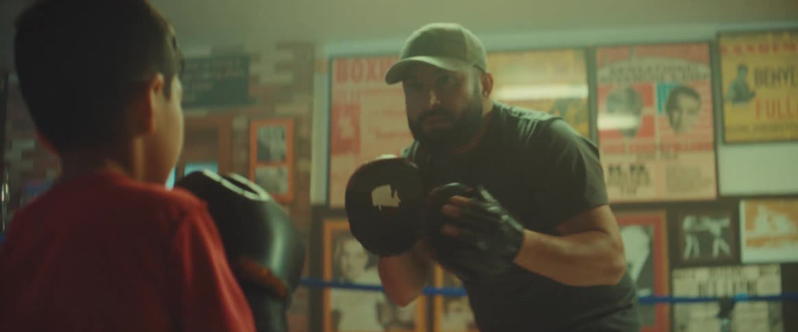 Man with cap black t shirt and boxing pads practices boxing with boy wearing red t shirt and boxing gloves in boxing ring
