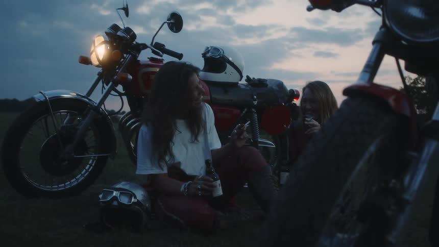 Two women sit on the grass near their motorcycles smoking and drinking beer