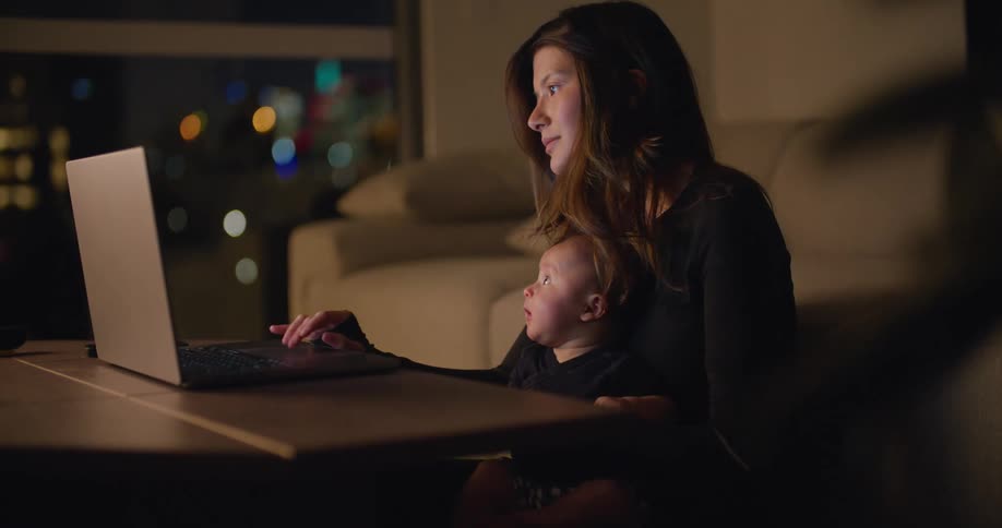 Mother holds and kisses the baby in her lap while using laptop