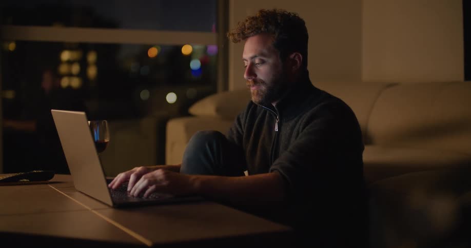 A man sits at a table drinking a glass of wine and typing on a laptop at night