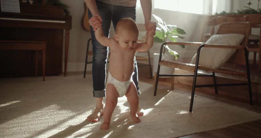 A woman helps a baby to walk across a living room during the day