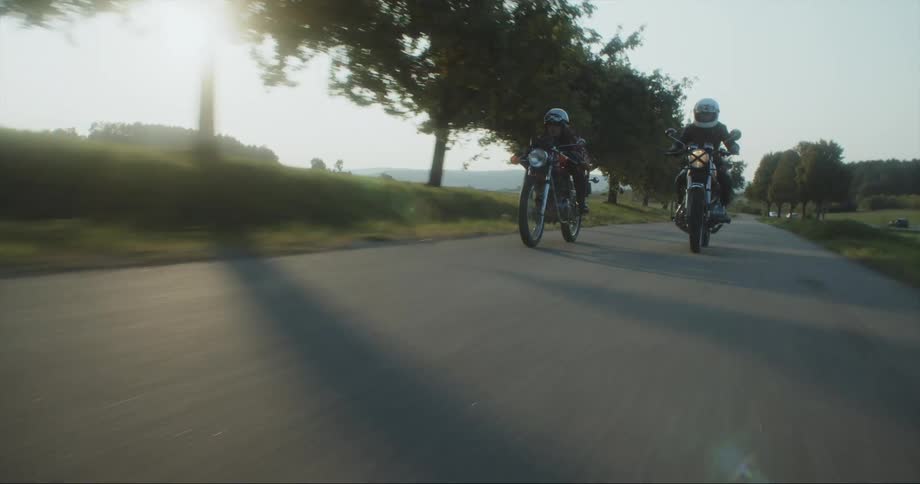 Two women on motorcycles zoom by a row of trees on a country road