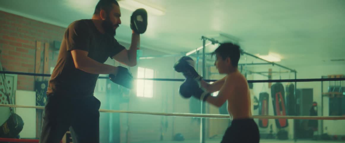 A boy practices boxing with a trainer in a boxing gym during the day