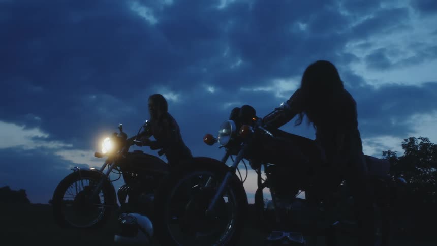 Two female bikers silhouetted against the sky start motorcycles before riding away at night
