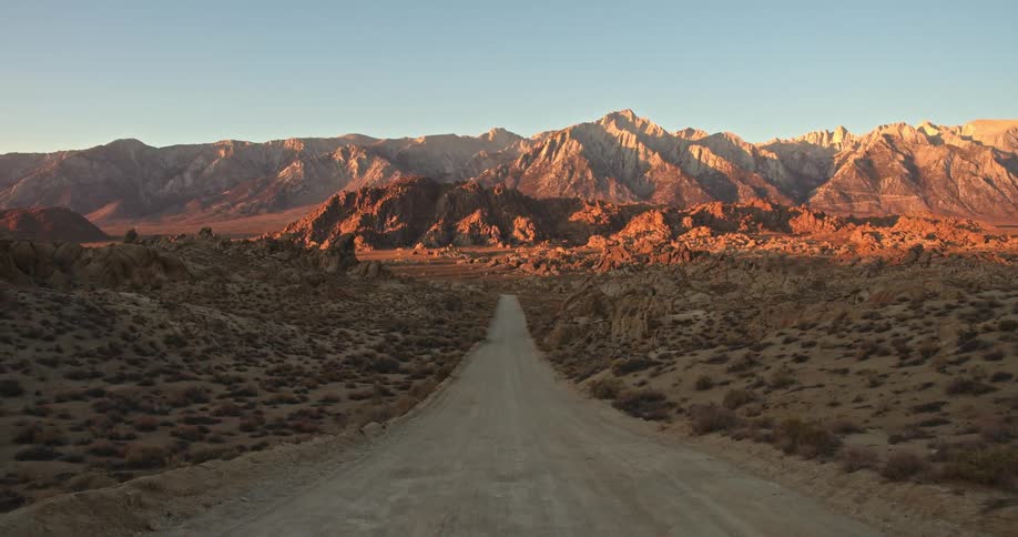 The sun shines on mountains at the end of a desert road under a clear blue sky