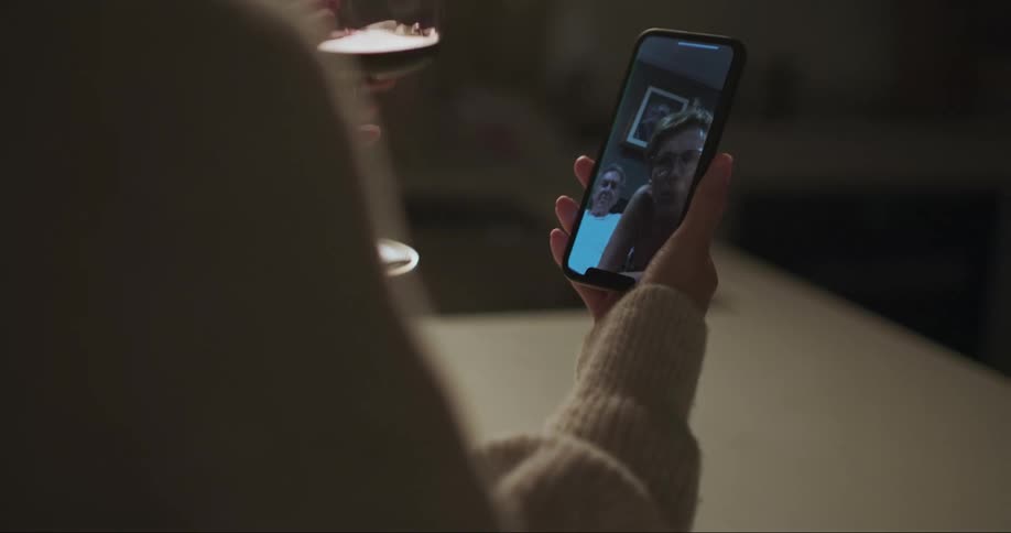A woman uses her smartphone to participate in a video call while holding a wine of glass