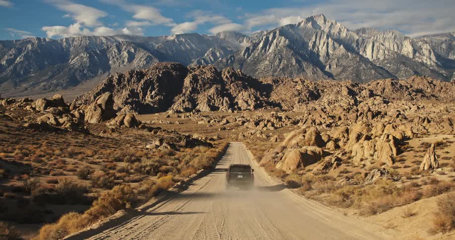 A truck travels on a desert road leading to mountains under a cloudy sky