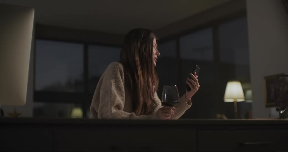 A woman speaks into her phone while holding a wineglass as she sits at a kitchen counter at night