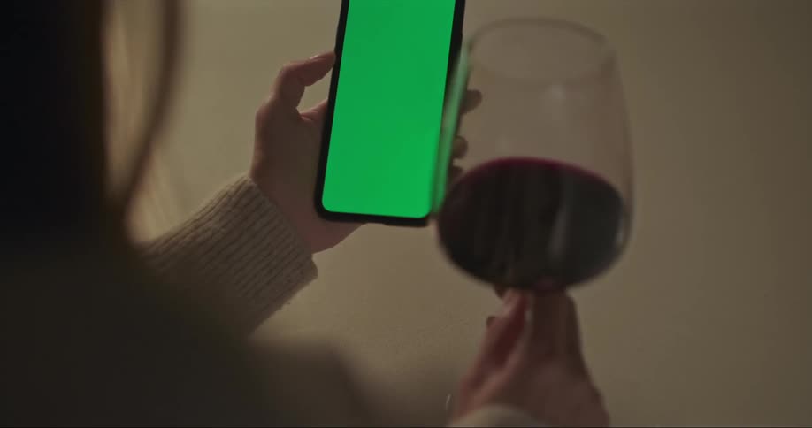 A woman is holding a cell phone with agreen screen in one hand and a glass of red wine in the other hand
