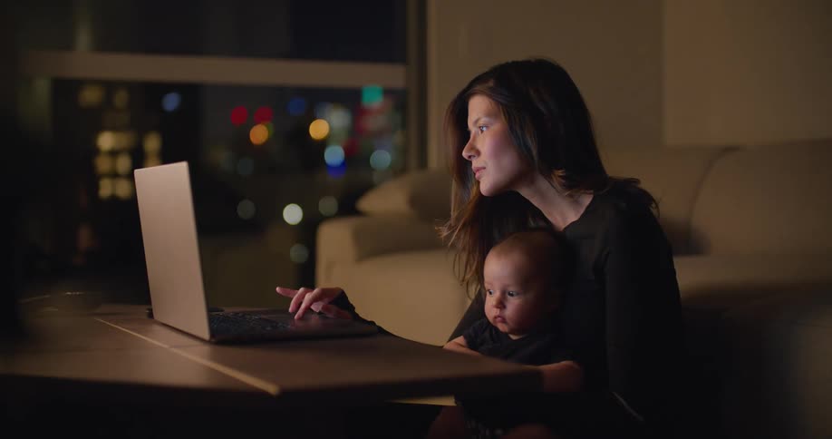 Woman with brown hair and black sweatshirt holds baby while using laptop in the living room