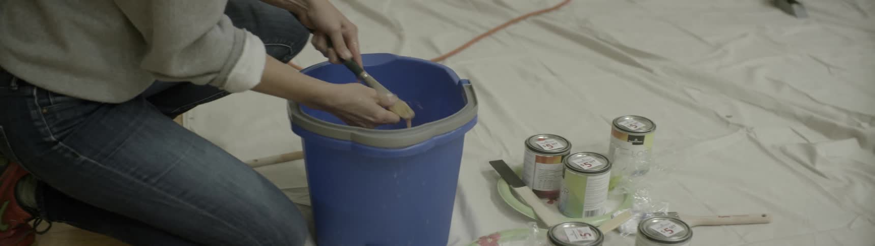 A woman kneeling down to clean her paint brushes in a blue bucket