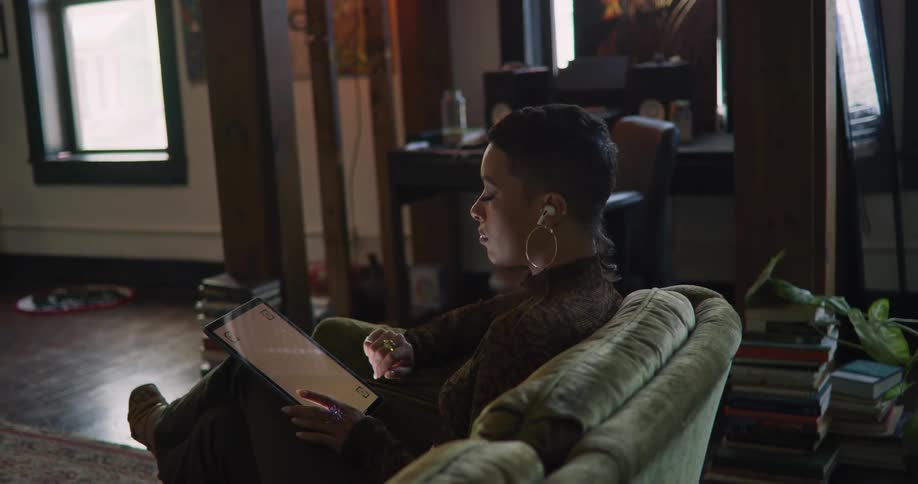 Woman with short hair hoop earrings and earphones sits on green couch using tablet