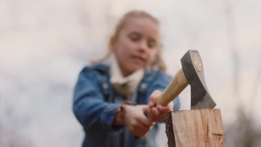Young blonde girl hits a piece of wood with an axe learning how to chop wood