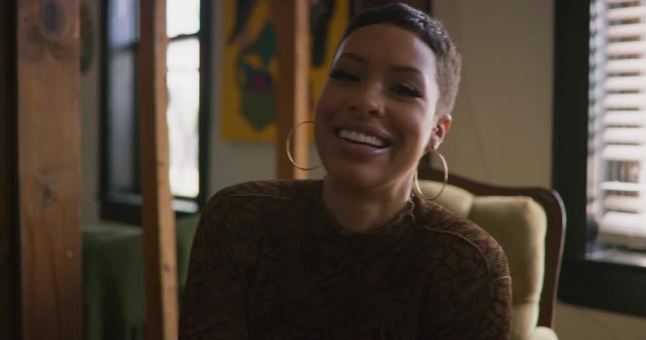 A young woman sitting indoors turns her face the camera while laughing
