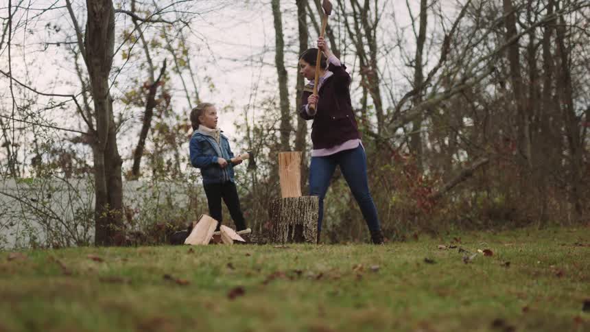 A woman teaches a girl how to chop wood outdoors on a cloudy autumn day