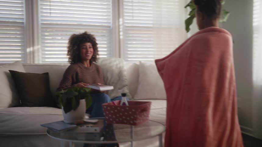 A woman puts down the book she is reading as she sits on the couch when her daughter walks into the room and hands something to her