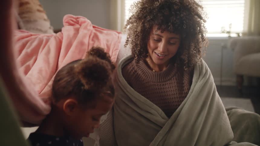 Mother bundled in blanket smiles and talks while watching daughter play