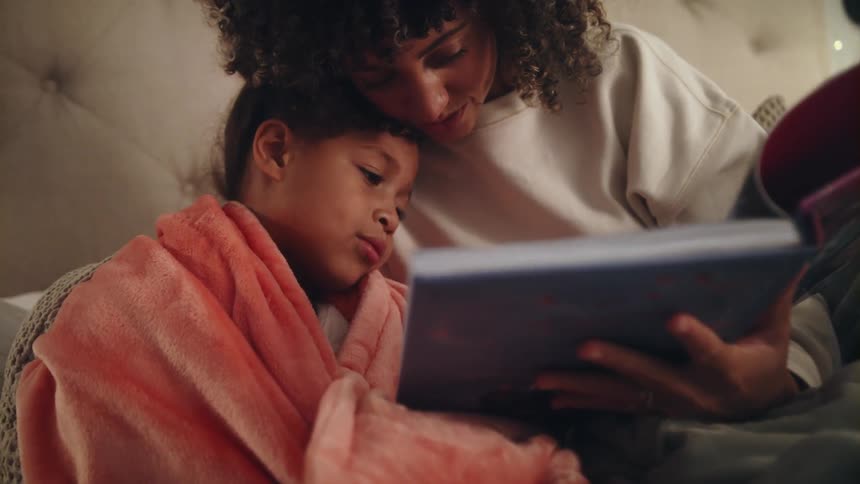 Mother with curly hair and beige sweatshirt reads book to daughter wrapped in blanket sitting on bed