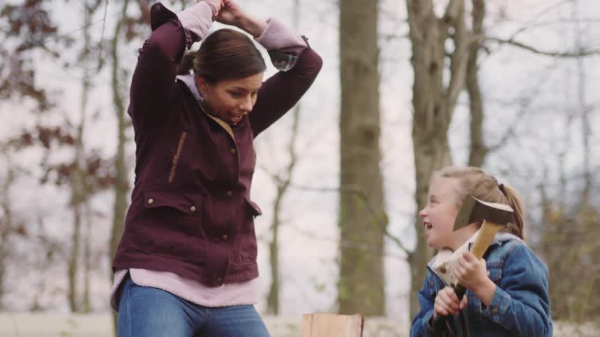 A little girl is standing outside holding an ax as her mother shows her how to split a piece of wood