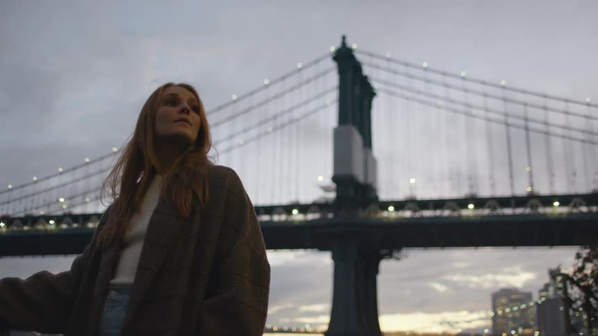 Long haired blonde woman standing in front of a suspension bridge in the city