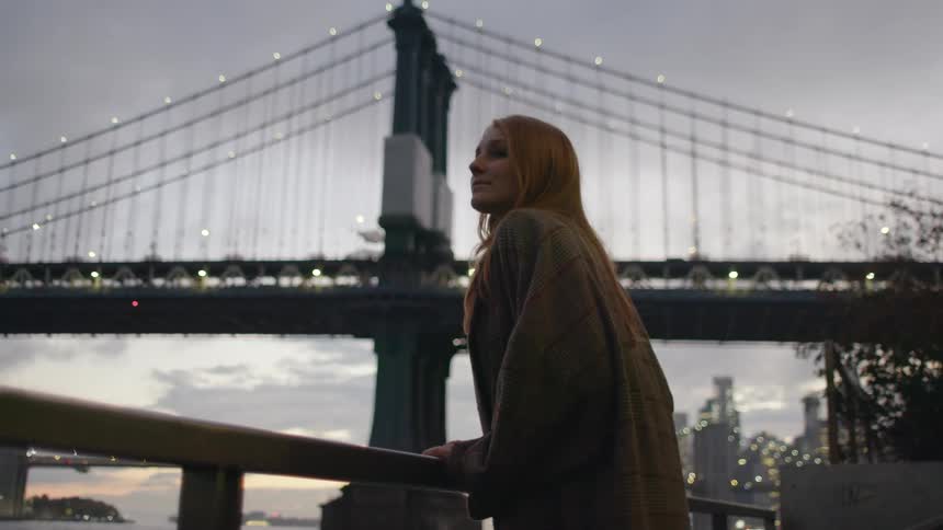 Young woman wearing a coat looks at the river under the new york bridges at evening