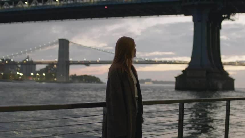 Young woman wearing a coat looking at a river under the bridges of new york