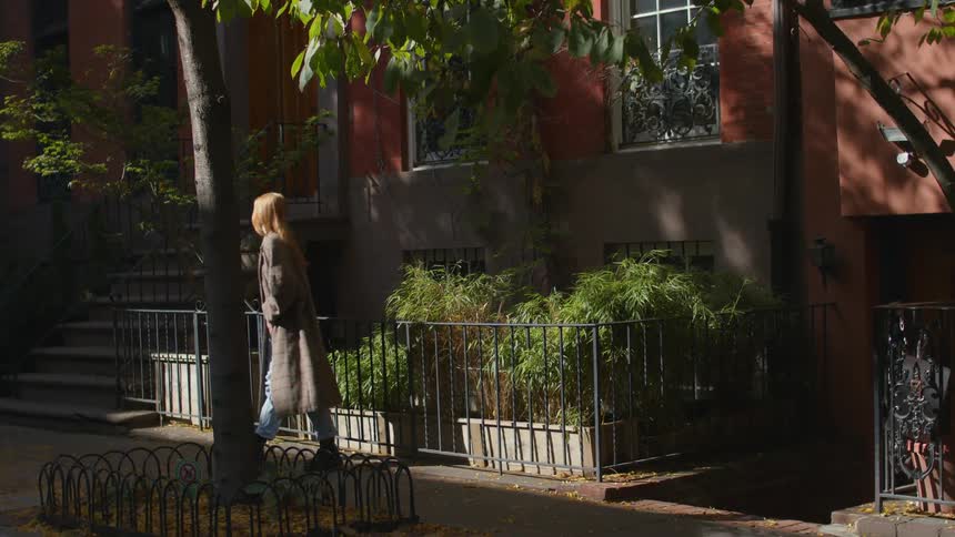 A woman reading a book while leaning against a fence outdoors closes her book and walks out of frame