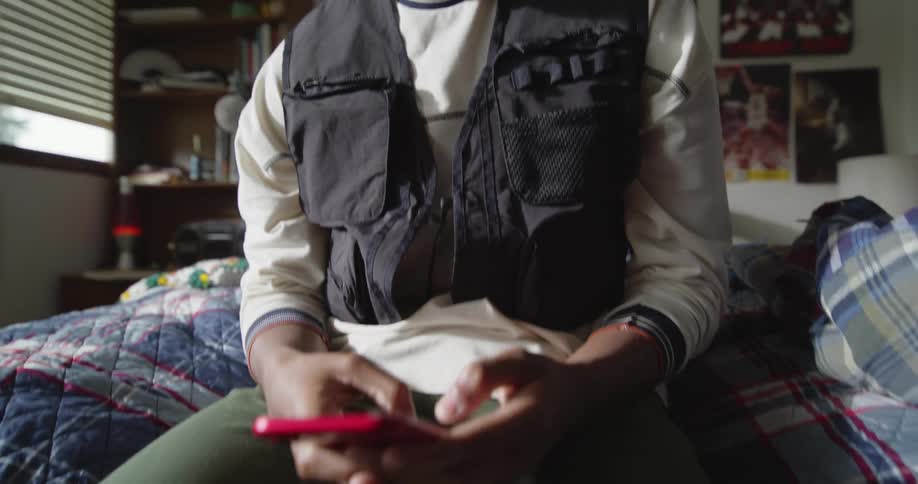 A young man uses his smartphone while sitting on a bed in a bedroom during the day