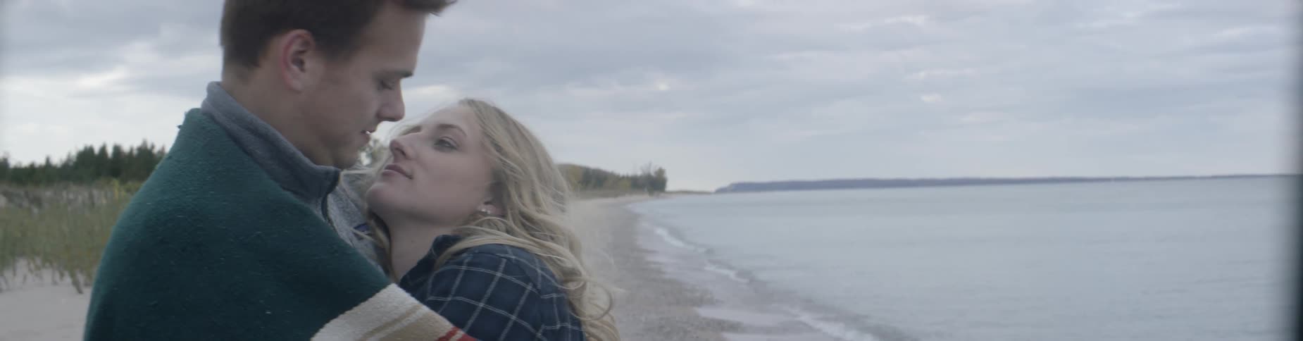 A man and woman cuddle and kiss on a beach on a cool overcast day