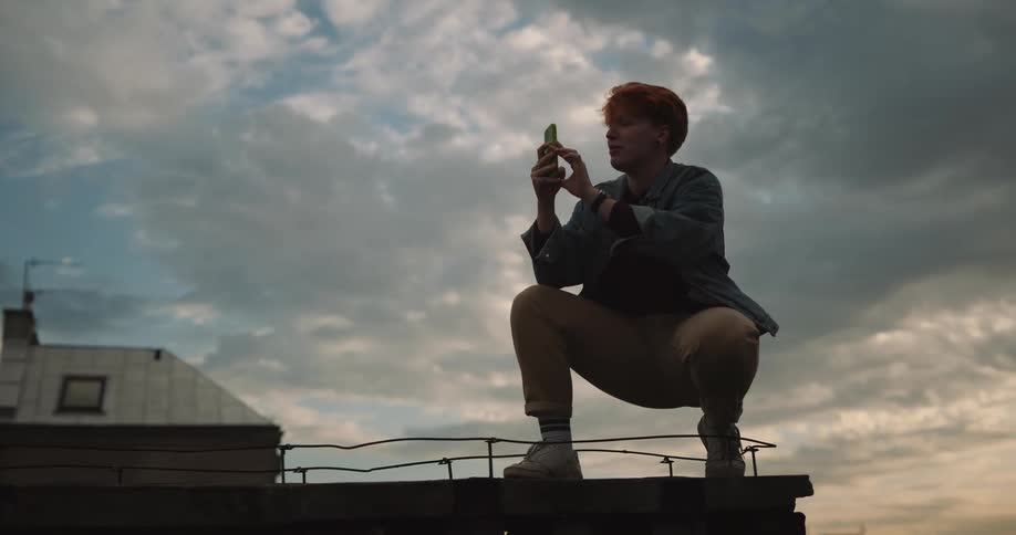 Woman crouches on roof and takes pictures using smartphone as another woman poses