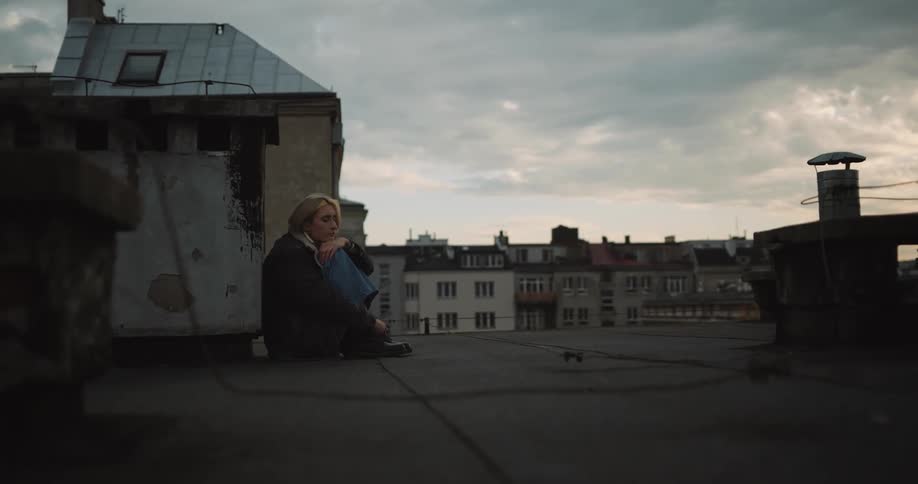 A woman hugs her knees while sitting on the roof of a building in a city on a cloudy day