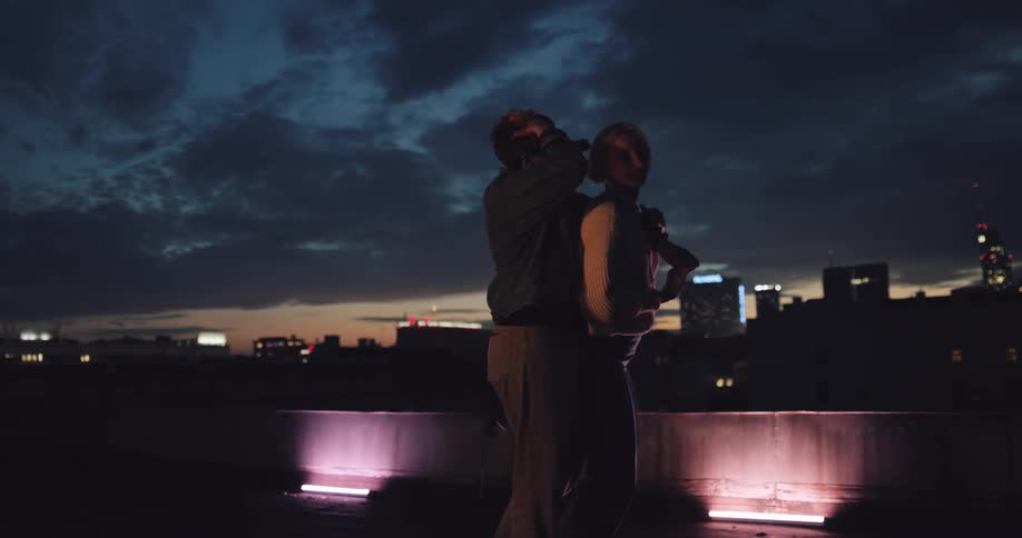 Two women slow dance and kiss on roof at night with city as backdrop