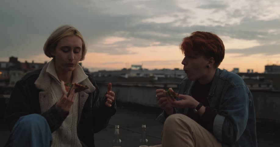 Two friends eating pizza on a rooftop at sunset