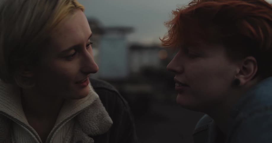 Woman with red hair kisses blonde woman outdoors at dusk