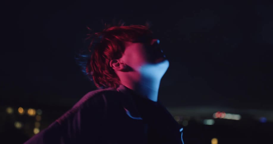 Redhead smokes and dances on roof at night with city lights in the background