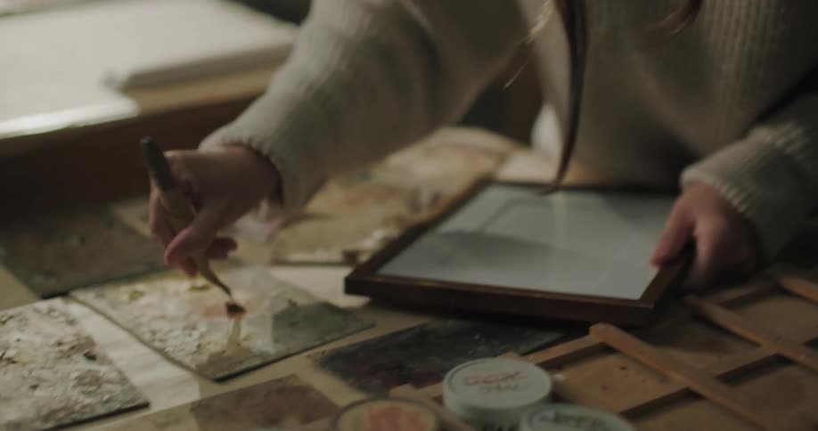 Woman picks up paint paste then applies it to frame while sitting at work table