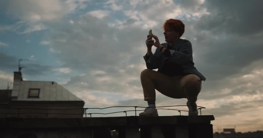 One person crouches and uses smartphone to take pictures as a female model takes different poses while they both stand on roof
