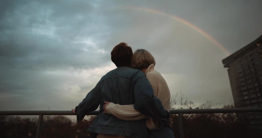 Two women hold hands as they walk across a roof top and look at a rainbow and then hug
