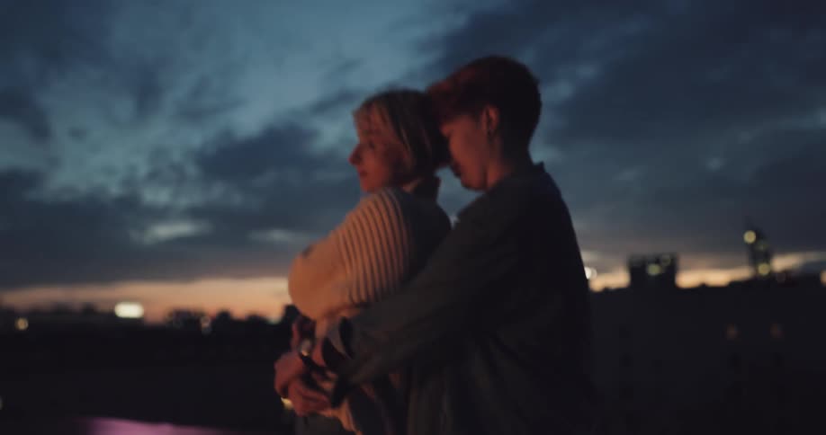 A man and woman are dancing and hugging outside near a lake with a city as the backdrop