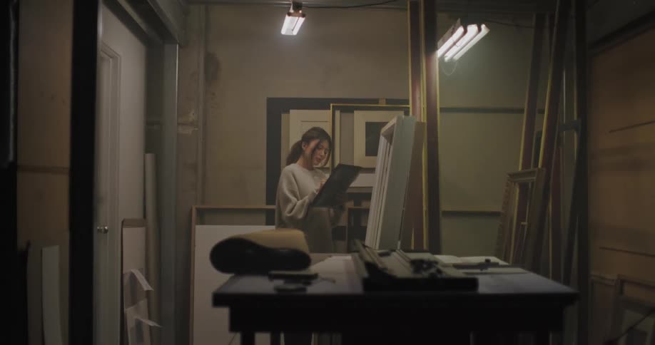 A woman in a warehouse flips through frames and writes on a paper on a clipboard