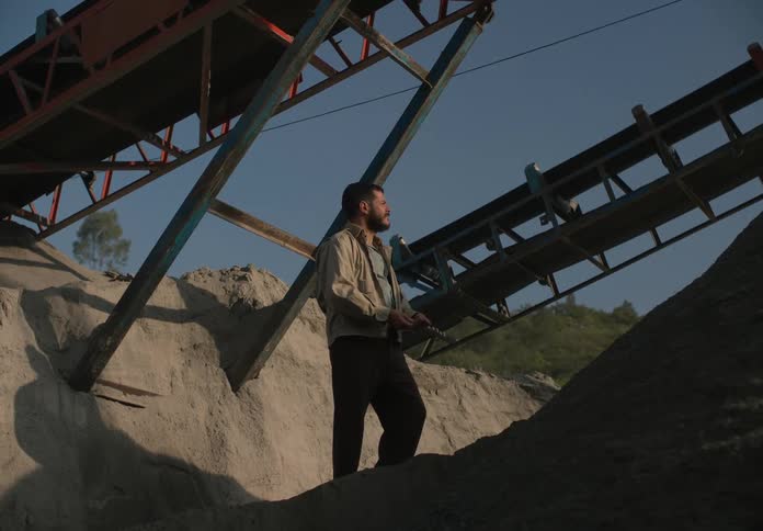 A man stands at a mining site and picks up some dirt and runs it through his fingers and looks around